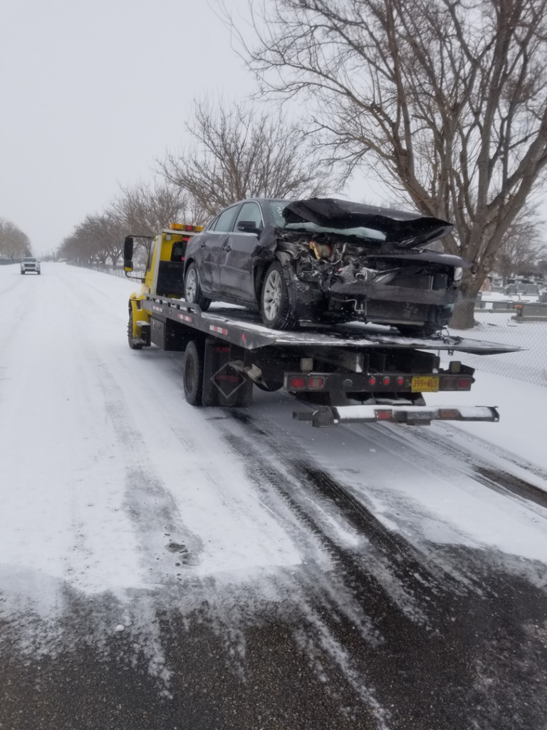 A flatbed tow truck from Gama Towing LLC transporting a damaged car on a snowy road in Carlsbad, NM.