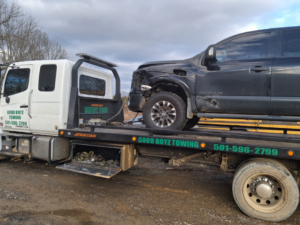 A flatbed tow truck from National Tow List hauling a damaged black pickup truck in Cove, AR.