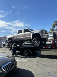 A flatbed tow truck hauling a large white pickup truck at Inglewood Transmission in Long Beach, CA.