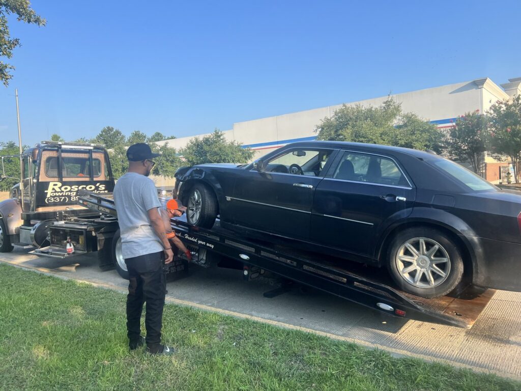 A flatbed tow truck from Rosco's Towing Services LLC loading a damaged black sedan in Carencro, LA.