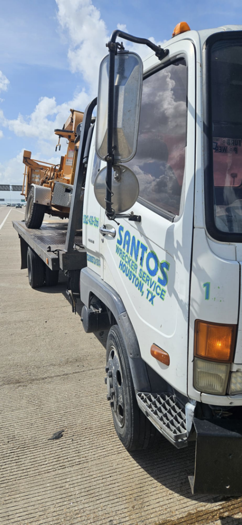 A flatbed tow truck from Santos Wrecker Services in Houston, TX, transporting a piece of yellow construction equipment.
