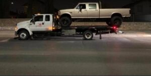A ROMU Towing flatbed truck actively towing a large pickup truck on a road at night in El Paso, TX.