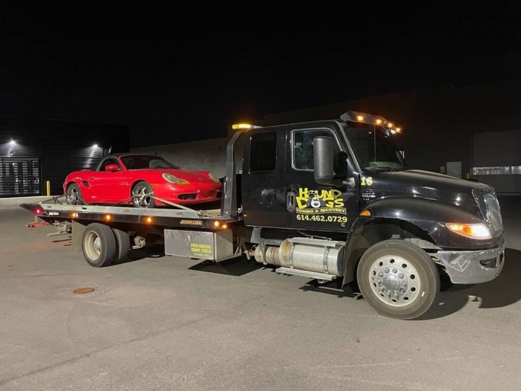 A Hound Dog's Towing & Recovery flatbed tow truck towing a red sports car at night in Columbus, OH.
