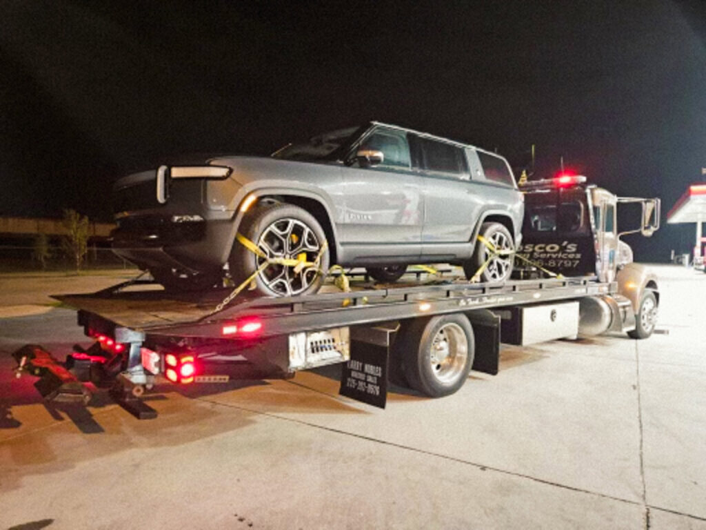 A flatbed tow truck from Rosco's Towing Services LLC towing a grey Rivian SUV at night in Carencro, LA.