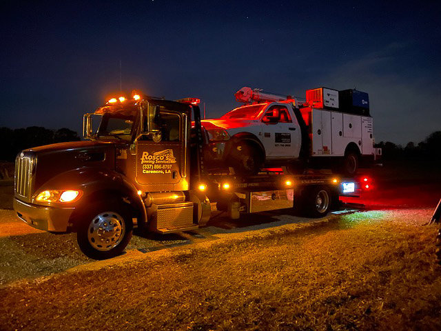 A flatbed tow truck from Rosco's Towing Services LLC towing a white service truck at night in Carencro, LA.