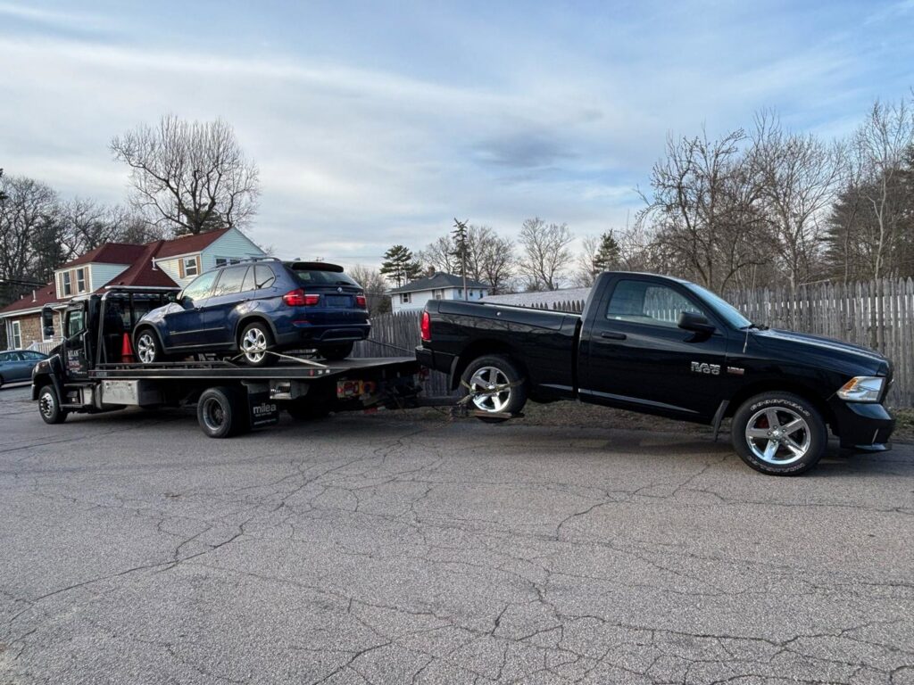 A Milan Towing inc. flatbed tow truck towing a blue SUV and a black pickup truck in Milan, MI.