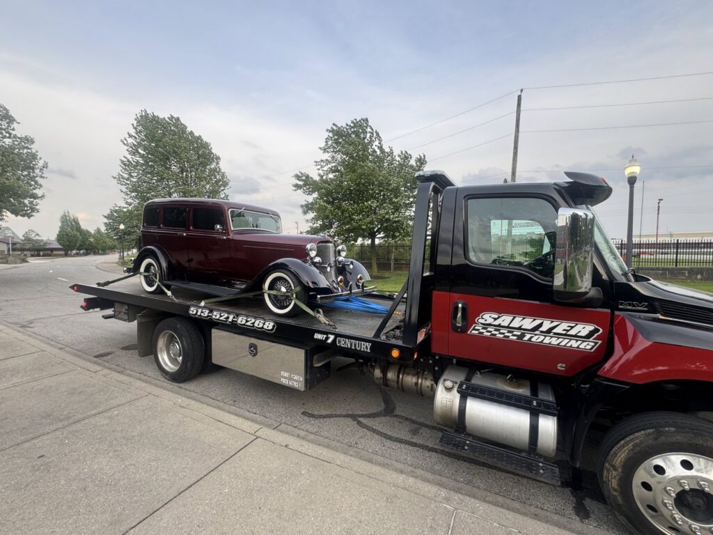 A vintage dark red car is securely loaded on a flatbed tow truck from Sawyer Towing in Cincinnati, OH.