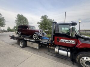 A vintage dark red car is securely loaded on a flatbed tow truck from Sawyer Towing in Cincinnati, OH.