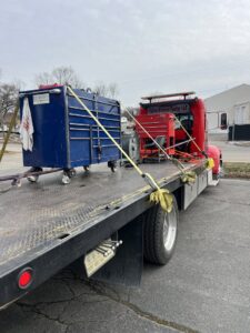 A red flatbed tow truck from Rubber Chicken Towing LLC transporting a large blue toolbox and other equipment in Charlotte, NC.