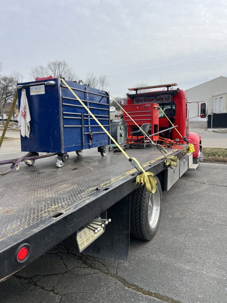 A red flatbed tow truck from Rubber Chicken Towing LLC transporting a large blue toolbox and other equipment in Charlotte, NC.