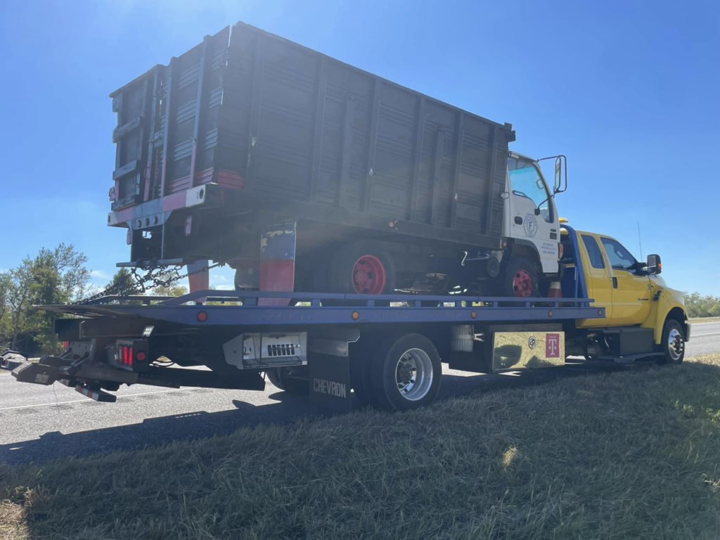 A yellow flatbed tow truck from Houston Towing transporting a white dump truck on its bed in Houston, TX.