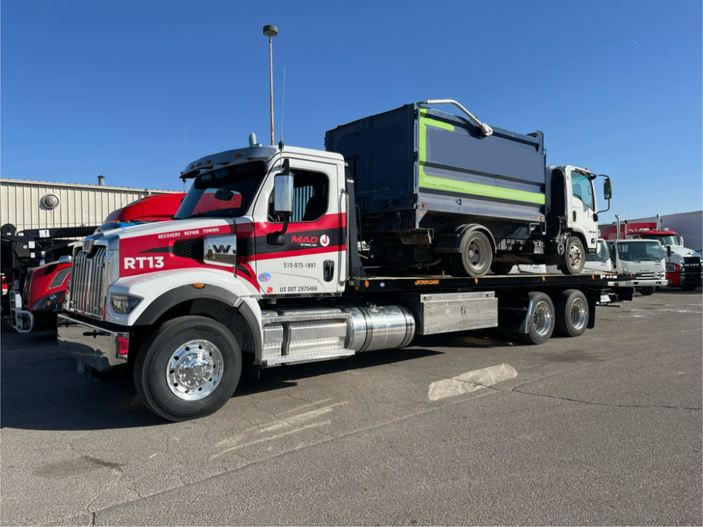 A flatbed tow truck from MAD J Repair & Towing transporting a smaller dump truck in Des Moines, IA.