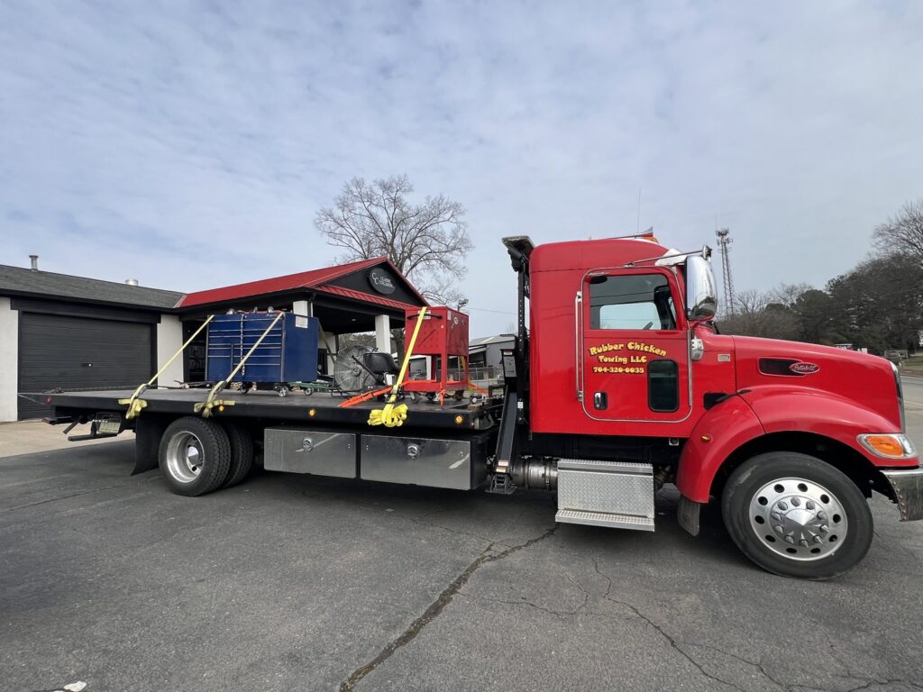 A red flatbed tow truck from Rubber Chicken Towing LLC transporting equipment, including toolboxes, in Charlotte, NC.