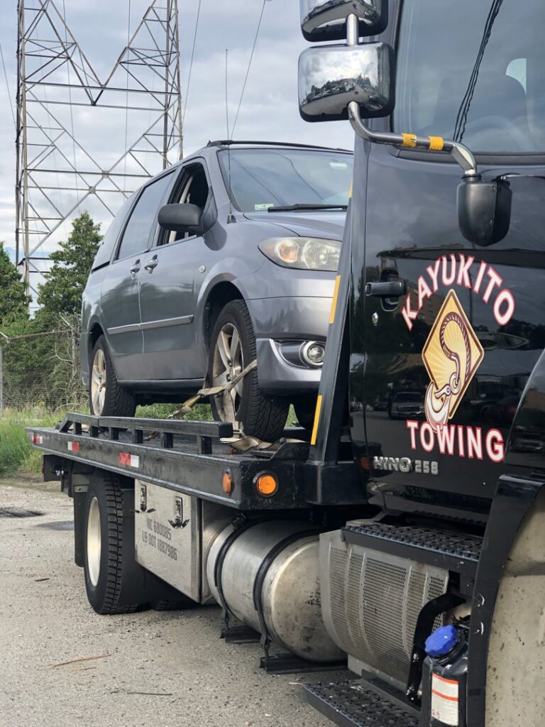 A flatbed tow truck from Cayuco Tires and Towing transporting a gray minivan in Worcester, MA.
