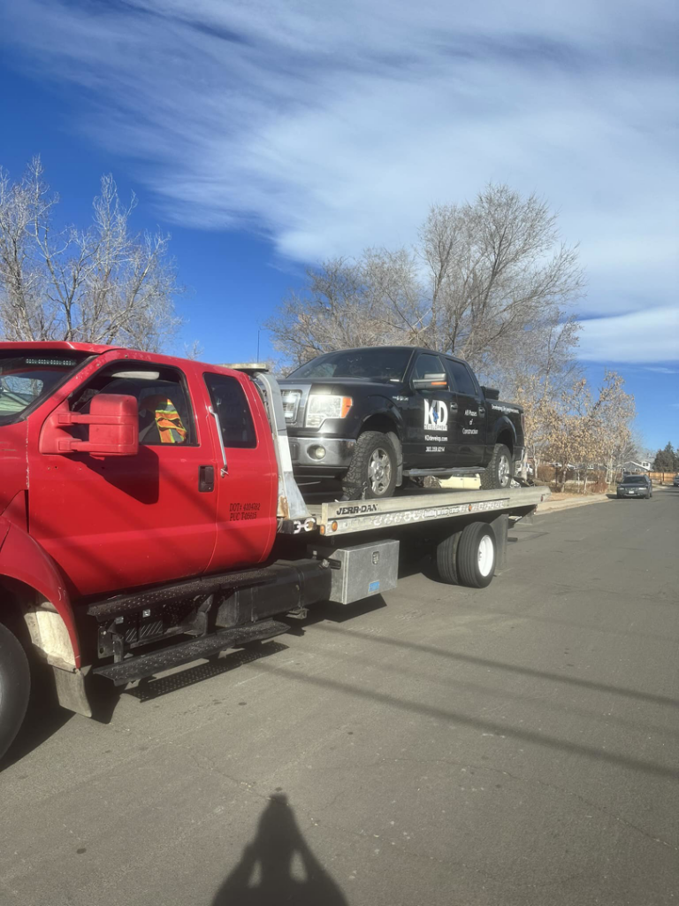 A red flatbed tow truck transporting a black pickup truck on a sunny day for Luck's Unlimited Towing in Thornton, CO.