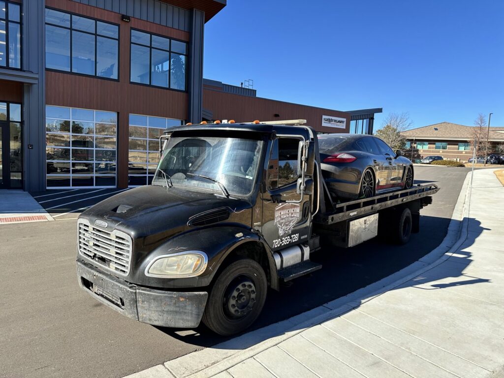 A flatbed tow truck from Henderson Towing & Recovery LLC transporting a dark grey Porsche Panamera in Denver, CO.