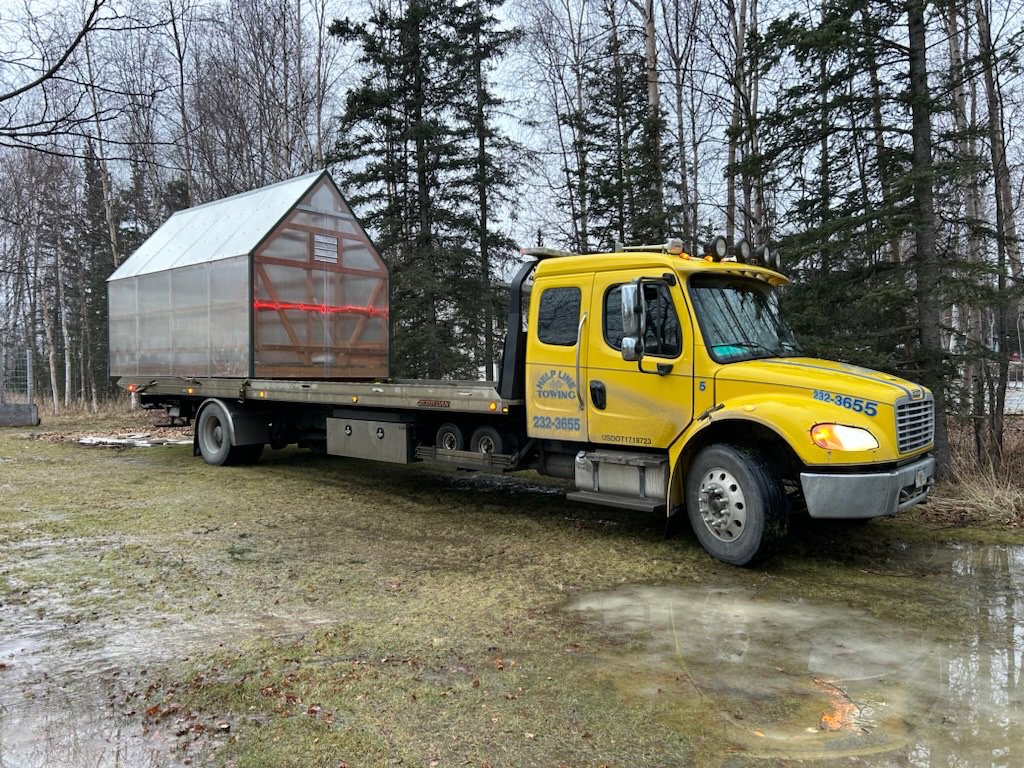 A Help Line Towing flatbed truck transporting a small shed or structure in a rural area of Wasilla, AK.