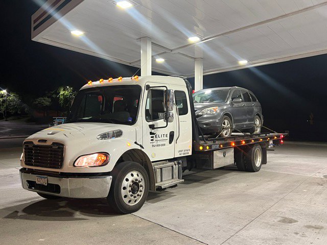 An Elite Towing of Minnesota flatbed tow truck with a grey car on its bed at a gas station at night in St Paul, MN.