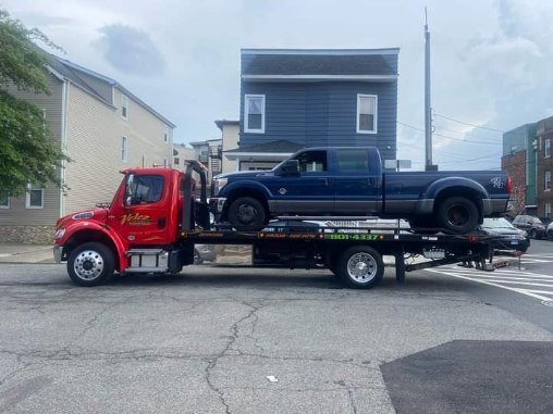 A Velez Towing flatbed truck transporting a blue pickup truck on a street in Newark, NJ.