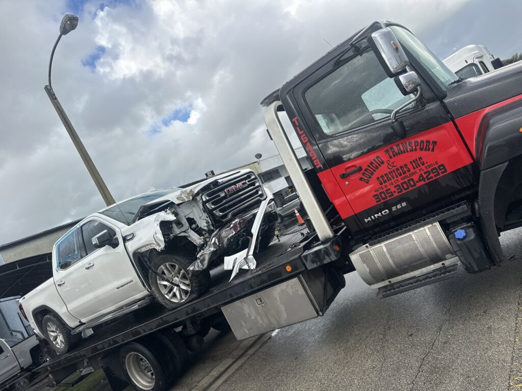 A flatbed tow truck from Rodicio Towing Services transporting a damaged white pickup truck in Hialeah, FL.