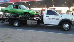 A white flatbed tow truck from All Pro Towing & Recovery, Inc. towing a green car at a gas station in Albuquerque, NM.