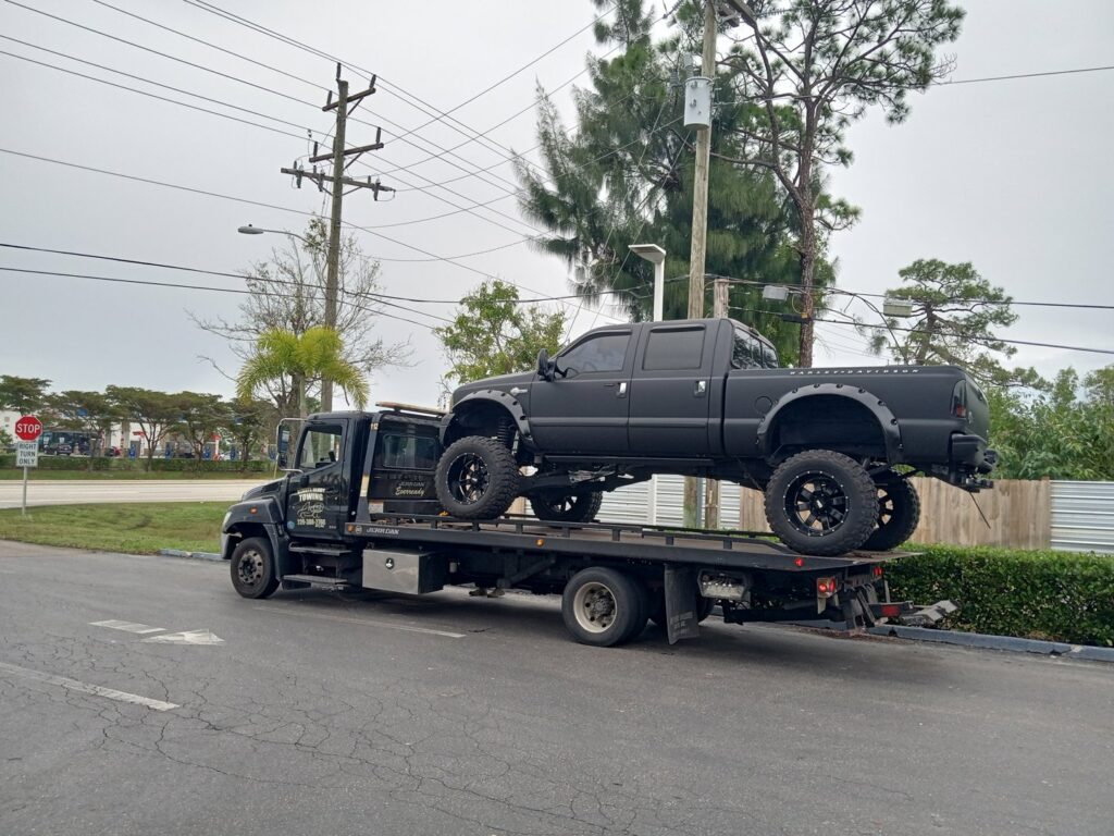 A flatbed tow truck from Always Ready Towing transporting a large black lifted pickup truck in North Fort Myers, FL.