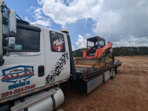 A flatbed tow truck from Allan Motors & Wrecker Service LLC towing an orange skid steer loader in Houston, TX