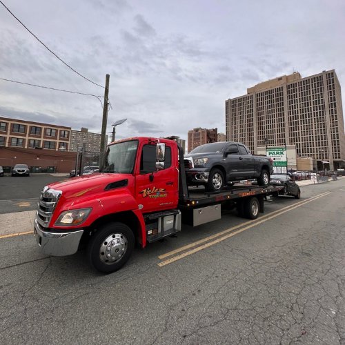 A Velez Towing flatbed truck transporting a grey pickup truck on a city street in Newark, NJ.