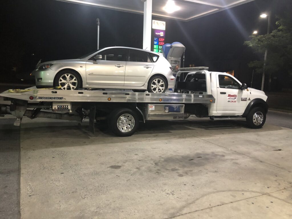 A white flatbed tow truck from Hank's Auto Service transporting a silver hatchback at night in Bethlehem, PA.