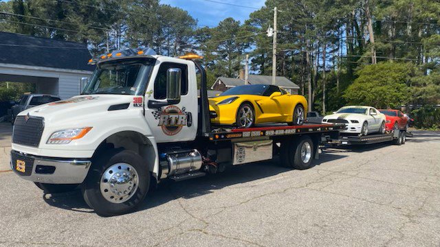 A flatbed tow truck from Raleigh Towing & Recovery transporting a yellow Corvette and a white Mustang in Raleigh, NC.