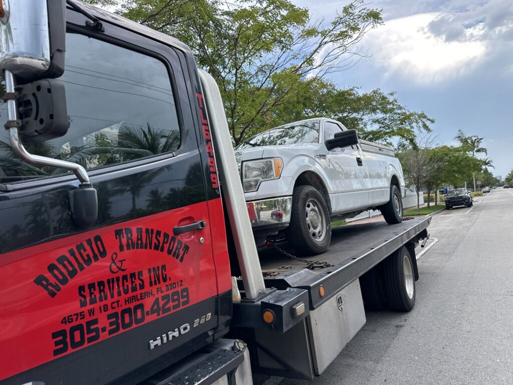 A flatbed tow truck from Rodicio Towing Services transporting a white pickup truck in Hialeah, FL.