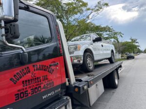 A flatbed tow truck from Rodicio Towing Services transporting a white pickup truck in Hialeah, FL.