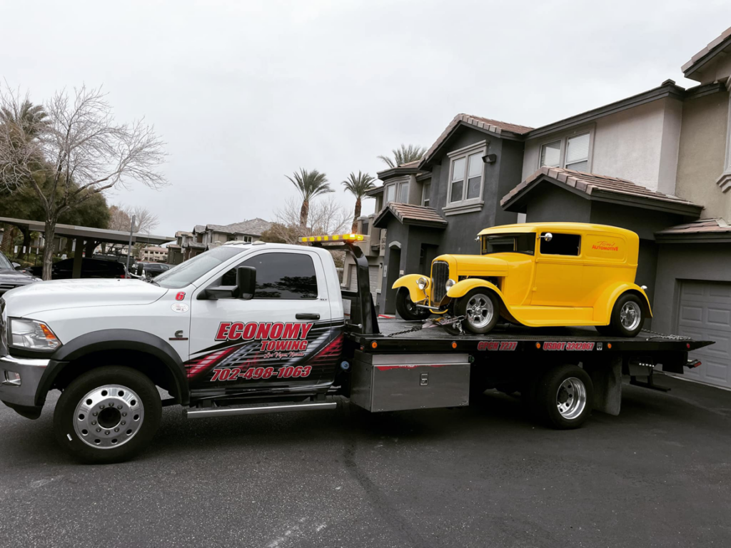 A white flatbed tow truck from Economy Towing transporting a yellow vintage car in Las Vegas, NV.