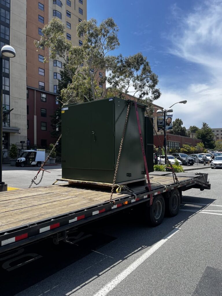 A flatbed trailer hauling a large green electrical box for Inglewood Transmission in Long Beach, CA.