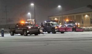 A fleet of three snow plow trucks parked in a snowy commercial lot at night, ready for service by Eric's Winter & Landscape Services in Appleton, WI
