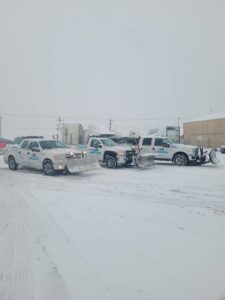 A fleet of JA Lawn Service, LLC trucks equipped with snow plows parked in a snowy lot in Fort Wayne, IN, ready for work.