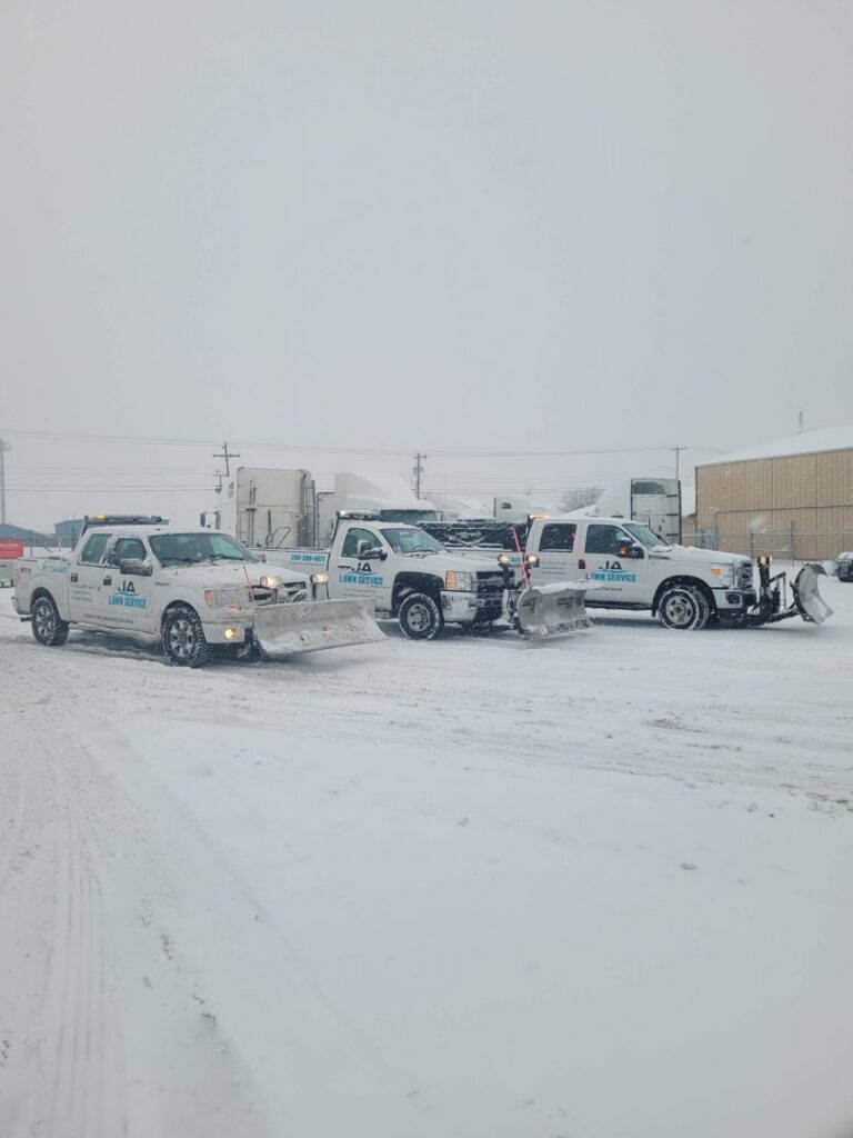 A fleet of JA Lawn Service, LLC trucks equipped with snow plows parked in a snowy lot in Fort Wayne, IN, ready for work.