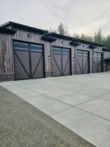 Four modern garage doors with dark frames and windows installed on a rustic-style building by Steelhead Doors, Services Available In.