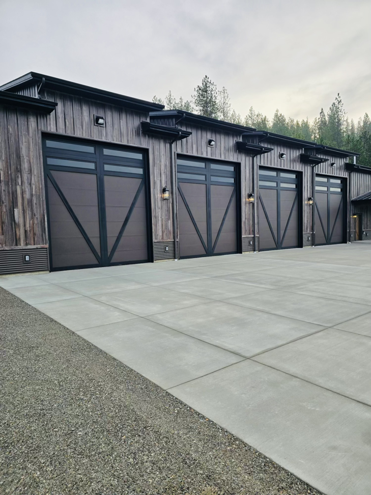 Four modern garage doors with dark frames and windows installed on a rustic-style building by Steelhead Doors, Services Available In.