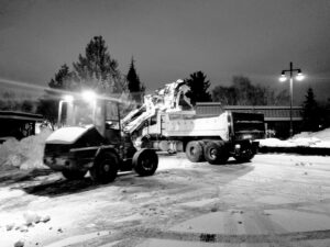 A front-end loader actively dumping snow into a large dump truck during a night snow removal operation by Yakima Landscape Management in Yakima, WA.