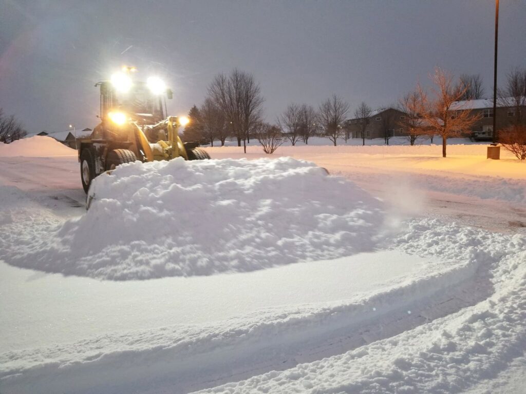 A front-end loader with a snow pusher attachment clearing a parking lot at night for Rochester Ground Lawn & Snow Services in Rochester, MN.
