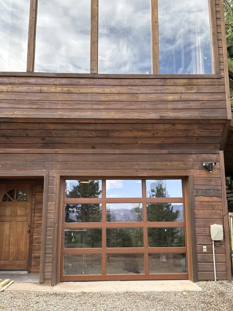 A modern wooden house featuring a stylish full-view glass garage door installed by Overhead Door Company of Santa Fe in Santa Fe, NM.