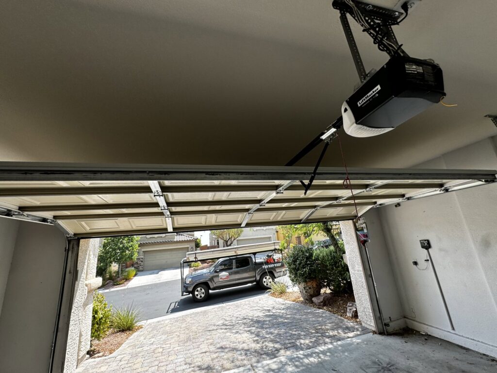 An interior view of a garage with the door partially open, showing the opener and tracks, with an Apex Garage Doors LLC truck outside in Las Vegas, NV
