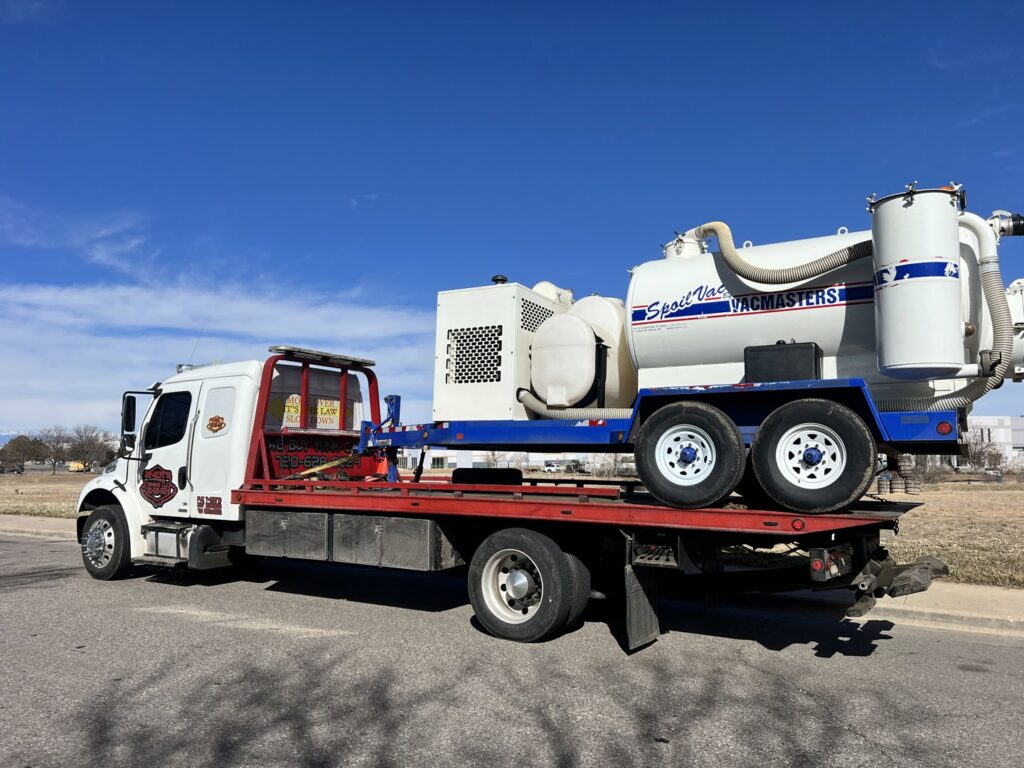 Giner's Towing flatbed truck transporting a large industrial vacuum trailer in Aurora, CO.
