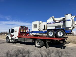 Giner's Towing flatbed truck transporting a large industrial vacuum trailer in Aurora, CO.