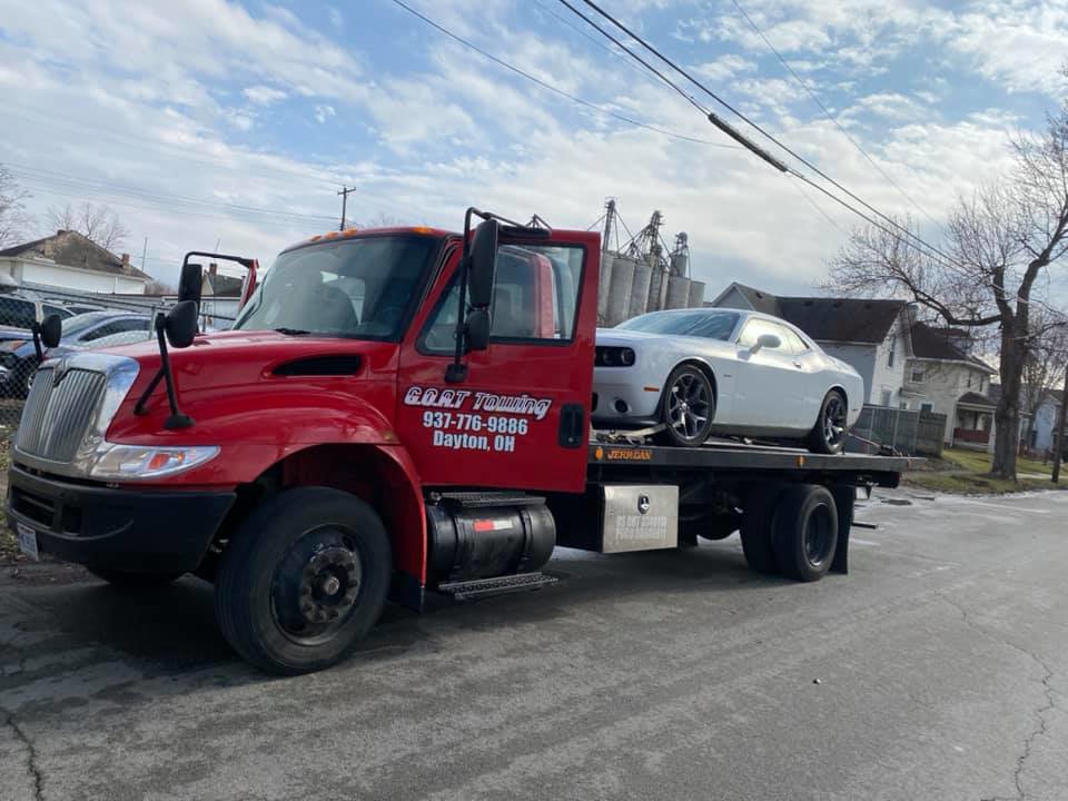 A red flatbed tow truck from Goat Towing transporting a white Dodge Challenger in Dayton, OH.
