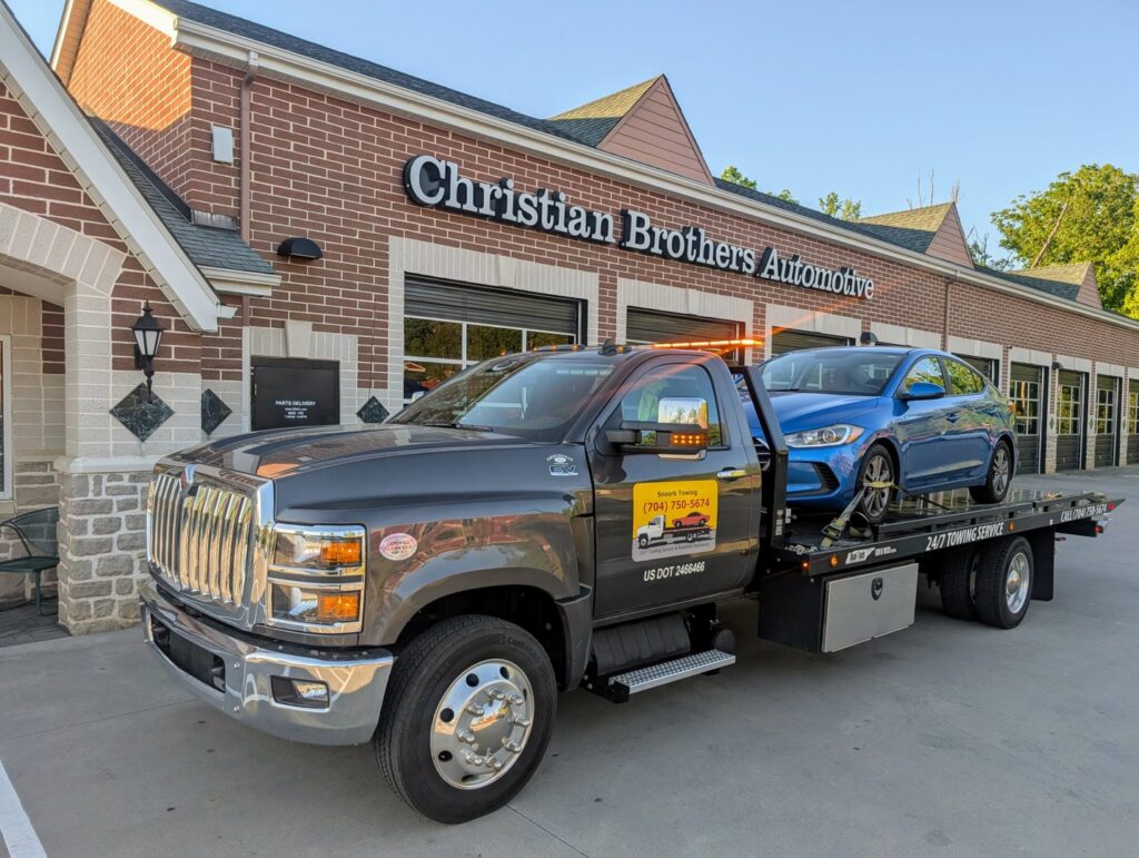 A gray flatbed tow truck from Snoork Towing transporting a blue sedan to Christian Brothers Automotive in Charlotte, NC.