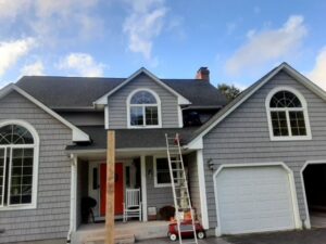 A house with newly installed gray shake siding, with a worker on the roof, by DL Roofing and Siding in New Haven, CT