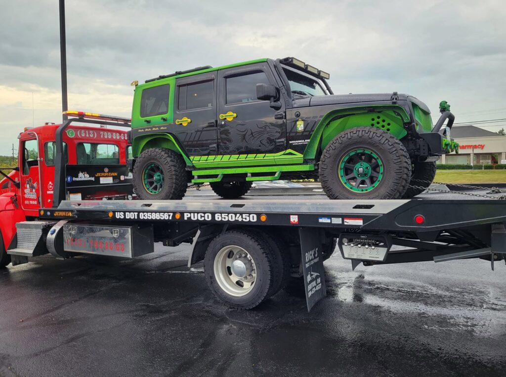 A green and black Jeep being transported on a flatbed tow truck by Vatos Locos Towing in Cincinnati, OH.