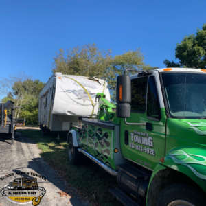 A green tow truck from Sunny Acres Towing towing a white travel trailer in Greenville, IN.